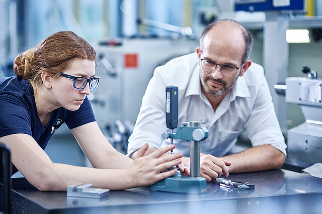 Azubine kontrolliert ein Drehteil unter Aufsicht des Ausbilders